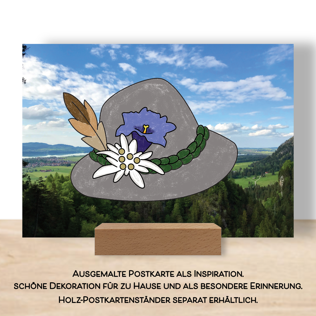 Ausgemalte Postkarte aus dem Postkartenset "Alpenzauber" als Beispiel. Aufgestellt im Postkarten Tischständer. Die Postkarte zeigt einen ausgemalten Trachtenhut mit Edelweiß, Enzian und einer Feder.