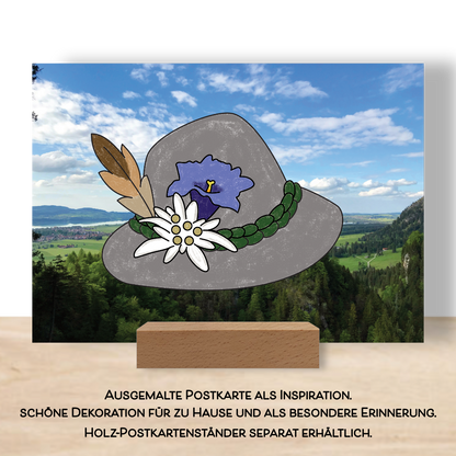 Ausgemalte Postkarte aus dem Postkartenset "Alpenzauber" als Beispiel. Aufgestellt im Postkarten Tischständer. Die Postkarte zeigt einen ausgemalten Trachtenhut mit Edelweiß, Enzian und einer Feder.