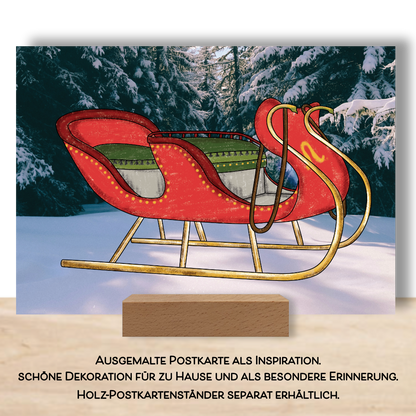 Ausgemalte Postkarte aus dem Postkarten Set "Weihnachtsglanz" Die Postkarte zeigt einen Schlitten der in rot und grün ausgemalt ist. Der Fotohintergrund zeigt eine winterliche Landschaft. Die Postkarte ist aufgestellt in einem Postkartenständer. 