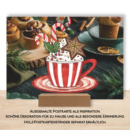 Ausgemalte weihnachtliche Postkarte als Inspiration im Postkartenständer aus dem Postkarten-Set "Glitzerzeit". Das Ausmalbild eine weihnachtliche Tasse ist ausgemalt in rot und weiß und verziert mit einem braunen Lebkuchen, rot weißen Zuckerstangen und einer grünen Mistel. 