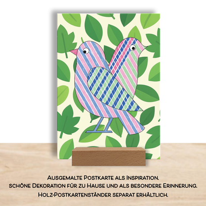 Fertig ausgemalte Postkarte aus dem Postkarten-Set "Color Pop". Es zeigt zwei Vögel auf einem Blätter-Fotohintergrund. Die Karte steckt in einem Holz-Tischständer für Postkarten. 