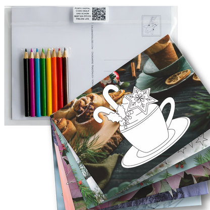 Weihnachtliches Postkarten-Set zum Ausmalen auf Fotohintergrund und 8 Buntstiften. Gezeigt werden  die weihnachtlichen Ausmalbilder gefächert und obenauf liegt eine Postkarte mit dem Ausmalbild einer Tasse mit Zuckerstangen, Stern und Mistelzweig. 